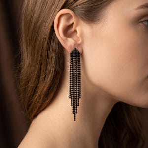 Peut inclure: Boucles d'oreilles pendantes noires composées de petits carrés. Les boucles d'oreilles sont fixées à l'oreille et pendent, créant un effet de frange. Les boucles d'oreilles sont de couleur foncée et le design est symétrique.