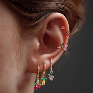 May include: A close-up of an ear adorned with multiple pieces of jewelry. Gold-toned hoop earrings dangle with cherry, pineapple, and grape charms. Other earrings include a strawberry, watermelon, and heart-shaped design, all with colorful gemstones.