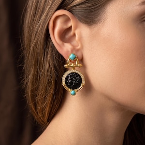 May include: A gold-tone drop earring featuring a central black circular cameo with a profile relief. The earring is accented with two small turquoise-colored stones, one at the top and one at the bottom, and a border of tiny clear crystals.