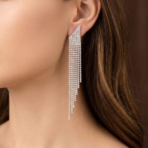 May include: A long, silver-toned dangle earring featuring a triangular top section encrusted with clear, sparkling rhinestones. Multiple delicate, vertical chains of varying lengths hang from the base, creating a cascading, stepped effect.