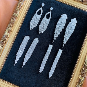 Op de afbeelding: Een assortiment zilveren oorbellen, gepresenteerd op een zwarte achtergrond in een gouden lijst. De oorbellen tonen diverse ontwerpen, waaronder druppel-, vierkante en lineaire stijlen, allemaal versierd met talrijke kleine, reflecterende stenen.