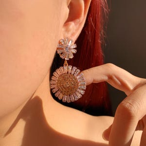 May include: Close-up of a gold-toned earring with a floral design. The earring features a smaller, starburst-shaped top and a larger, circular bottom, both adorned with clear, sparkling crystals. The earring is worn on a person's ear.