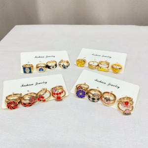 May include: A collection of fashion jewelry rings displayed on white cards. The rings feature various designs, including evil eyes, bows, and geometric patterns. The rings are gold-toned with colorful enamel accents.