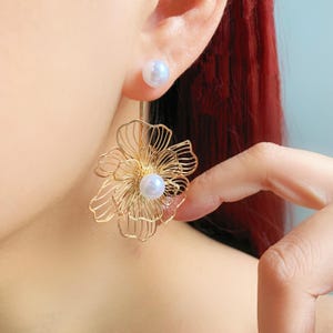 Puede incluir: Pendientes florales dorados con un perno de perla y una perla más grande en el centro del diseño floral. Los pendientes tienen un diseño calado delicado y se muestran en una oreja.
