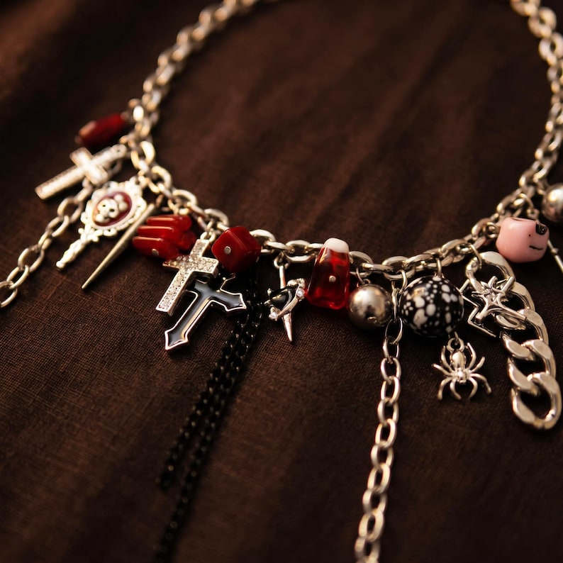K&ouml;nnte beinhalten: Silberfarbene Halskette mit verschiedenen Anh&auml;ngern, darunter Kreuze, rote Perlen, eine Spinne und ein rosa Schwein. Der dunkle, braune Hintergrund hebt die metallischen und farbigen Elemente hervor.
