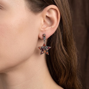 Peut inclure: Une boucle d'oreille pendante en métal foncé avec un pendentif en forme d'étoile orné de petites pierres précieuses facettées rouges et orange. La boucle d'oreille possède une attache à clou et un élément en étoile avec une finition métallique texturée.