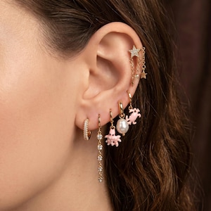 May include: A collection of gold-toned earrings worn in a single ear. The set includes a star-shaped stud with dangling chains, a small hoop with a pearl, a hoop with a pink octopus charm, a hoop with a pink crab charm, and two hoops with clear stones.