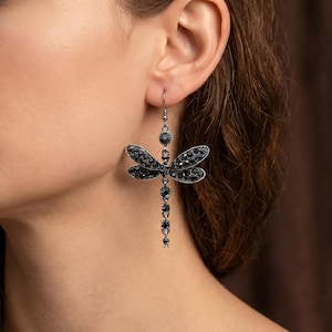 Può includere: Orecchino pendente con un design a forma di libellula. La libellula è adornata con gemme nere, con una gemma circolare in cima e una serie di gemme che si estendono verso il basso. L'orecchino è argentato e ha una chiusura a gancio.