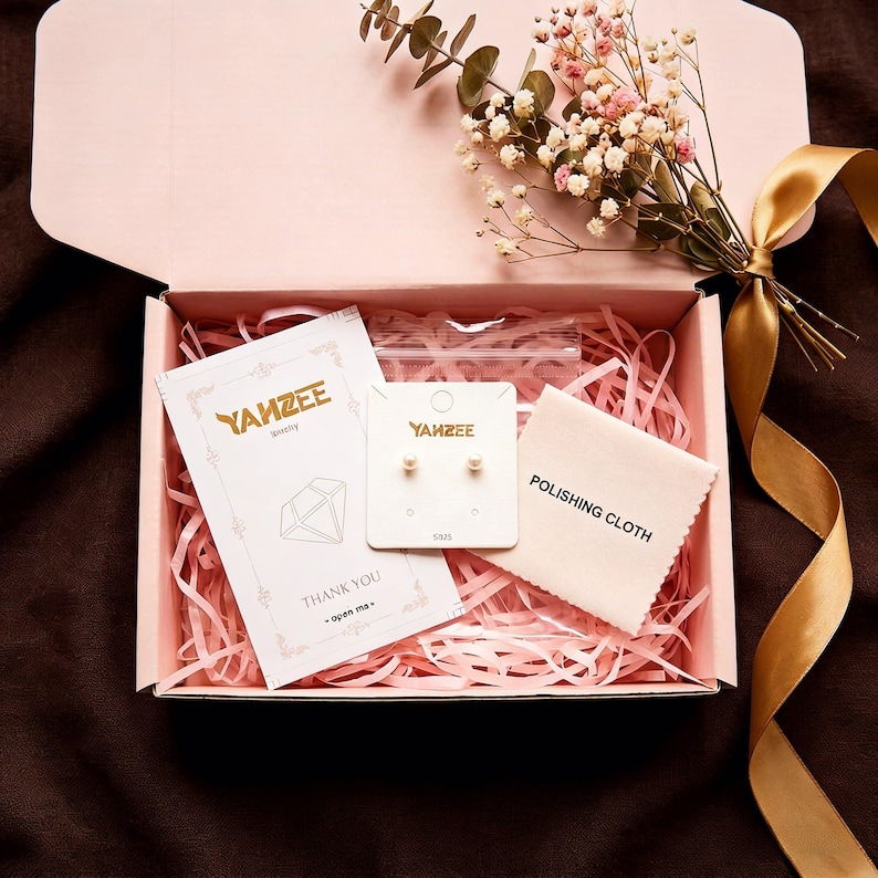 K&ouml;nnte beinhalten: Eine rosa Geschenkbox mit goldenem Band, die Schmuck und Accessoires enth&auml;lt. Darin befinden sich ein Paar Perlenohrringe auf einer Karte, ein Poliertuch und eine Dankeskarte. Die Box ist mit rosa Papierstreuseln gef&uuml;llt und mit Blumen verziert.