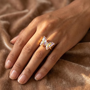 May include: A gold-colored ring featuring two iridescent butterfly wings with a small, clear gem in the center. The ring is worn on a finger, showcasing the intricate details and shimmering effect of the butterfly design.