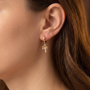 May include: A gold hoop earring featuring a small dangling palm tree charm. The palm tree pendant is crafted from gold-toned metal and is accented with a vertical row of tiny, clear, sparkling stones set along the center of the tree trunk.