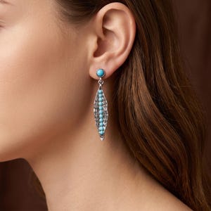 May include: A silver drop earring featuring a single round turquoise bead at the top and a textured, elongated silver pod shape below, which contains a vertical row of eight small, round turquoise beads set within the centre of the metal.