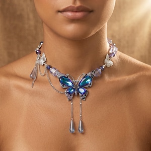 Op de afbeelding: Een close-up van een vlinderketting. De ketting heeft een grote, blauwe en paarse kristallen vlinderhanger. De ketting heeft zilveren kettingen en kleinere paarse en blauwe kralen. Twee druppelvormige kristallen hangen aan de vlinder.