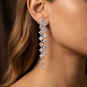 Pode incluir: Brincos pendurados elegantes com uma série de pedras preciosas claras de corte quadrado. Os brincos são cravejados em metal prateado e caem em cascata em um design linear. As pedras preciosas refletem a luz, criando um efeito brilhante.