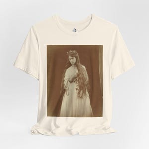 May include: Off-white t-shirt displaying a sepia-toned image of a woman in a long white dress. The woman has long, flowing hair, a floral crown, and a beaded necklace. The image has a vintage aesthetic, reminiscent of an old photograph.
