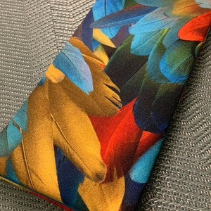 May include: A rectangular clutch purse with a colorful feather print design. The feathers are in shades of blue, red, yellow, and brown.