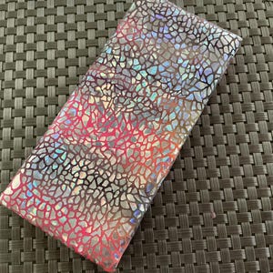 Puede incluir: Una pieza de tela rectangular con una base de color rosa vibrante cubierta por un patrón reflectante e iridiscente de vidrio agrietado. La superficie muestra un brillo metálico multicolor que cambia de apariencia a través de las formas geométricas.