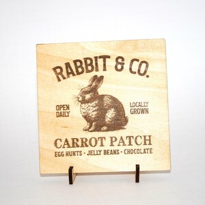May include: A wooden sign with the text "RABBIT & CO." and a drawing of a rabbit. The sign also says "OPEN DAILY", "LOCALLY GROWN", and "CARROT PATCH EGG HUNTS - JELLY BEANS - CHOCOLATE".