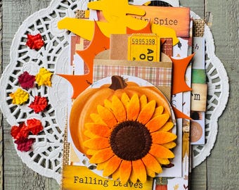 May include: A layered autumn-themed craft project featuring a large orange sunflower and pumpkin. The design includes paper cutouts of leaves, a yellow silhouette, and the text "Falling Leaves" and "in Spice". The arrangement is set on a white doily.