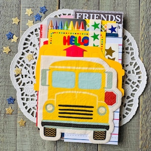 May include: A collection of school-themed stationery items, including a yellow school bus-shaped napkin, crayons, and stickers. The word "HELLO" is visible on a yellow card. The items are arranged on a white lace doily, with star-shaped confetti scattered around.