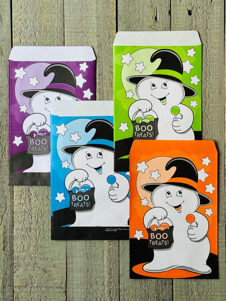 Pu&ograve; includere: Quattro sacchetti di dolcetti di Halloween con un fantasma che indossa un cappello da strega che tiene un lecca-lecca e una borsa che dice "Boo Treats!" I sacchetti sono viola, verde, blu e arancio.