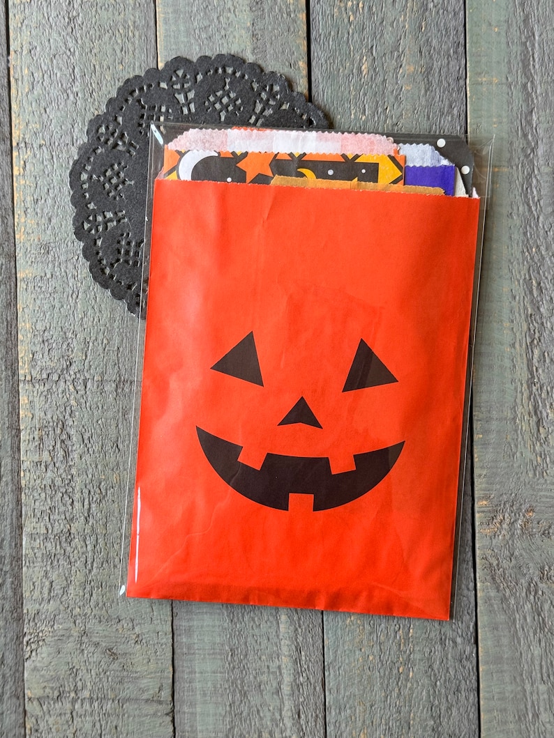 Pu&ograve; includere: Un sacchetto di carta rosso con una faccia di zucca nera stampata su di esso. Il sacchetto &egrave; pieno di ritagli di carta colorati e con motivi.
