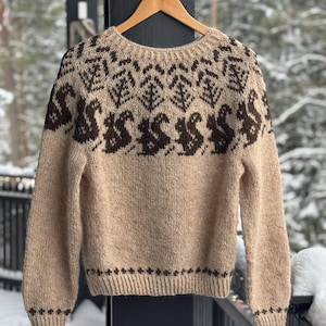 Puede incluir: Un suéter beige tejido con cuello redondo y mangas largas. El suéter presenta un patrón repetido de ardillas y árboles marrones en la pechera. Los puños y el dobladillo están detallados con un borde marrón. El suéter está colgado en una percha de madera.