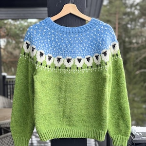 May include: A knitted jumper in green and blue, featuring a sheep design. The top section is light blue with white dots, while the lower part is green. A row of sheep with black faces and white bodies adorns the jumper.