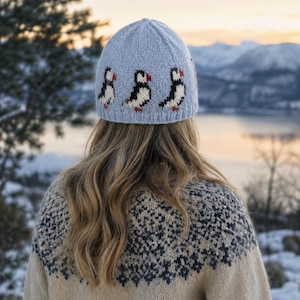 Puede incluir: Un gorro de punto azul claro con un patrón repetitivo de frailecillos negros, blancos y rojos alrededor de la base. El gorro tiene un borde acanalado y está confeccionado con un material de hilo suave, mostrando un diseño detallado de las aves.