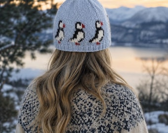 May include: A light blue knitted beanie featuring a repeating pattern of black, white, and red puffin birds around the base. The hat has a ribbed edge and is constructed from a soft yarn material, displaying a detailed intarsia design of the birds.