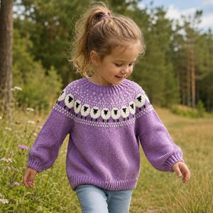 May include: A lavender long-sleeve knit sweater featuring a round neckline and a yoke decorated with a repeating pattern of white and black sheep. The garment has ribbed cuffs and a ribbed hem, constructed with a soft, textured wool-like yarn.