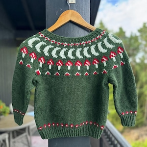 May include: A dark green long-sleeve sweater featuring a round neckline with a knit pattern of light green crescent moons and red and white mushrooms. The cuffs and hem are ribbed with a small red and white geometric pattern above the trim.