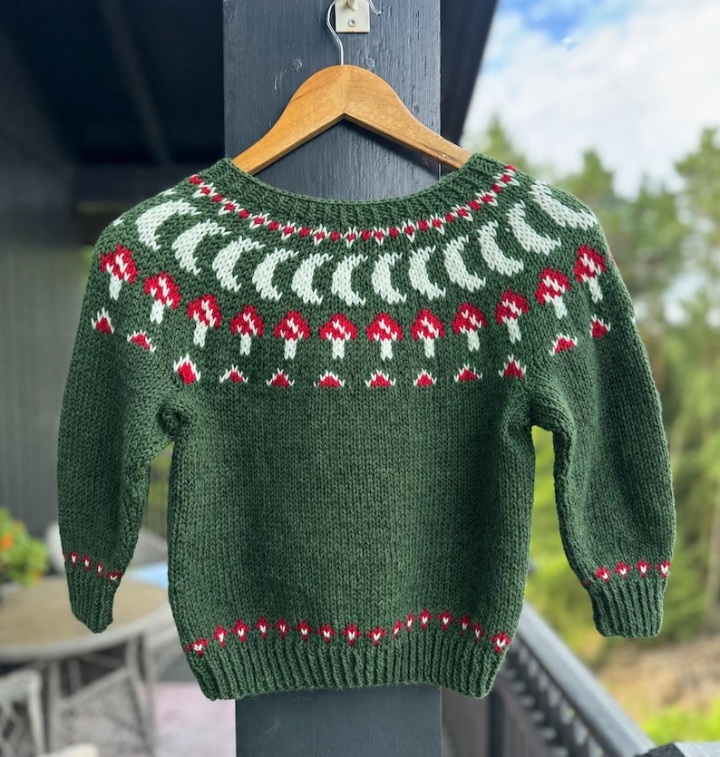 May include: A dark green long-sleeve sweater featuring a round neckline with a knit pattern of light green crescent moons and red and white mushrooms. The cuffs and hem are ribbed with a small red and white geometric pattern above the trim.