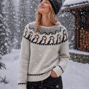 Pode incluir: Um suéter de manga comprida na cor creme com um padrão fair isle em cinza escuro e preto com motivos repetidos de papagaios-do-mar ao redor do decote, combinado com um gorro de malha preto e jeans cinza escuro num dia de inverno com neve.