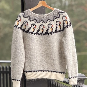 May include: A light gray knit sweater with a crew neck, featuring a repeating pattern of puffin birds across the chest. The sweater has a white bottom hem and cuffs, with black detailing. The sweater is hanging on a wooden hanger.
