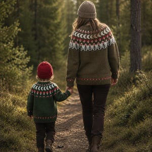 May include: Two individuals wearing matching olive green knit sweaters with a white and red patterned yoke featuring crescent moons and mushroom motifs. The taller individual wears a tan beanie and dark pants, while the smaller wears a red beanie.