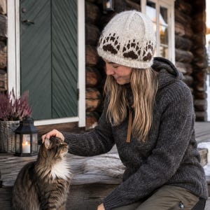 Könnte beinhalten: Eine cremefarbene Strickmütze mit einem sich wiederholenden dunkelbraunen Bärenpfoten-Muster am unteren Rand. Die Mütze ist aus einem strukturierten, wolleartigen Garn gefertigt und hat eine abgerundete Form für einen bequemen Sitz.