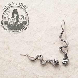 Può includere: Un paio di orecchini a forma di serpente color argento con squame testurizzate e chiusura a gancio. L'immagine include un logo circolare con una persona in posa meditativa e il testo ALMA LIBRE by Eva Kohlschitter.