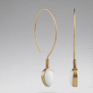 Può includere: Un paio di orecchini color oro con un lungo gancio curvo che si trasforma in una sezione metallica a spirale. Ogni orecchino termina con una pietra bianca liscia a forma di goccia, offrendo un aspetto pulito ed equilibrato.