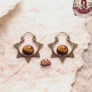 Può includere: Un paio di orecchini a cerchio color bronzo con una montatura a forma di stella e una pietra preziosa occhio di tigre rotonda al centro. Un piccolo ciondolo a forma di fiore di loto in legno intagliato è posto tra i due.