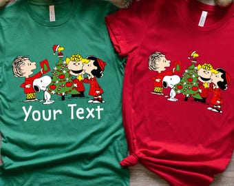 May include: A green t-shirt and a red t-shirt, each featuring a graphic of the Peanuts characters decorating a Christmas tree. The green shirt includes the white text Your Text below the graphic. Both shirts have short sleeves and crew necklines.