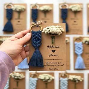 Op de afbeelding: Marineblauwe macramé sleutelhanger met een kwast, bevestigd aan een gouden ring, weergegeven op een bruine kaart. De kaart heeft de namen "Maria" en "Jason" met een hartsymbool, en de tekst "Bedankt voor het vieren met ons!" en de datum "02.05.2022".