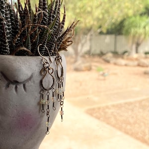 以下が含まれることがあります： 手作りのブロンズ色のイヤリング。揺れるデザインで、複雑なワイヤーワークと小さな虹色のビーズが特徴です。白い顔型のプランターに多肉植物と一緒に飾られています。