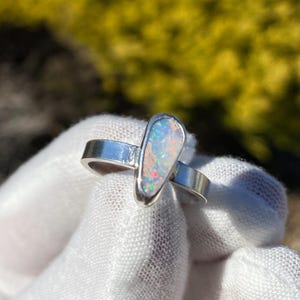 May include: A silver ring featuring a teardrop-shaped opal gemstone set in a smooth metal bezel. The opal displays iridescent flashes of red, blue, and green colour play against a light, semi-translucent base within a polished silver band.