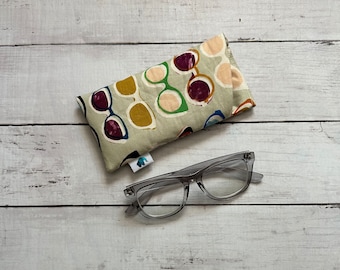 May include: Clear-framed spectacles and a soft eyeglasses case. The case is beige with a repeating pattern of sunglasses in various colours, including purple, green, and yellow. The spectacles are rectangular.