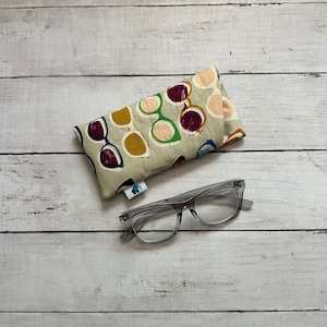 May include: A pair of clear-framed eyeglasses and a soft eyeglasses case. The case is beige with a repeating pattern of sunglasses in various colors, including purple, green, and yellow. The eyeglasses are rectangular.