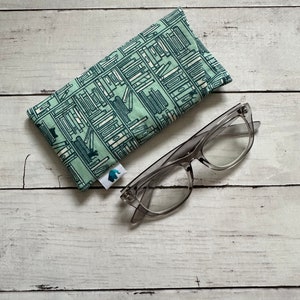 May include: A green and white eyeglass case with a bookshelf pattern. The case is closed and lying on a white wooden surface. A pair of clear glasses is lying next to the case.