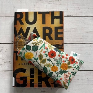 May include: A floral print glasses case with a book titled "Ruth Ware The Woman in Cabin 10" on a white wooden surface. The glasses case features a cream background with red, yellow, and green floral patterns. The book has a brown and black cover.