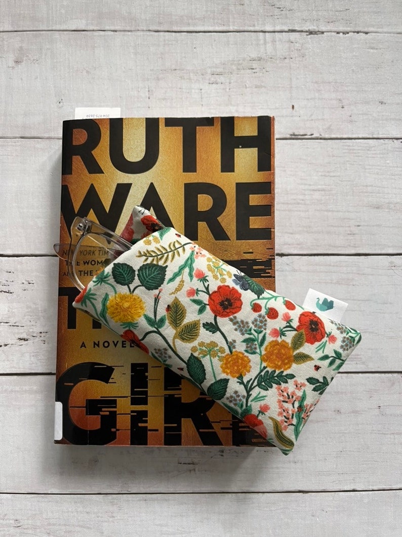 May include: A floral print glasses case with a book titled "Ruth Ware The Woman in Cabin 10" on a white wooden surface. The glasses case features a cream background with red, yellow, and green floral patterns. The book has a brown and black cover.