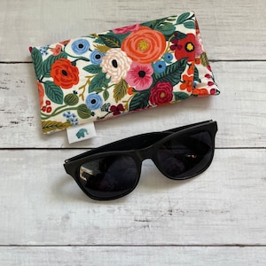 May include: A floral patterned eyeglass case with a white background and colourful flowers. The case is lying on a white wooden surface next to a pair of black sunglasses.
